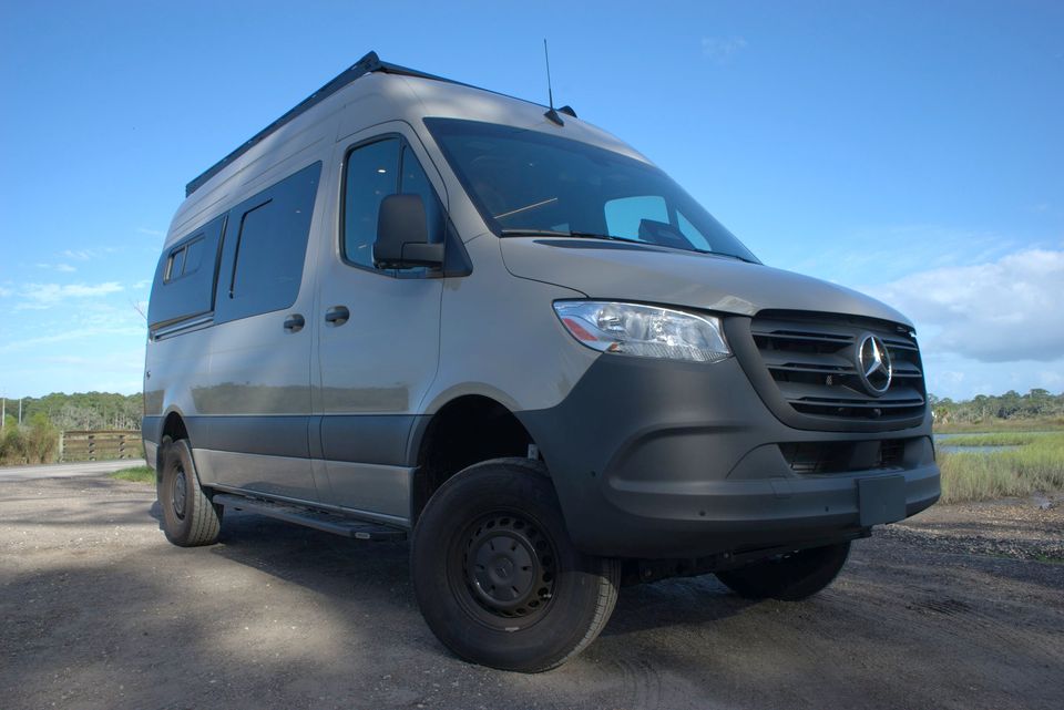 A gray Drifter camper van with black wheels and a roof rack on a gravel lot