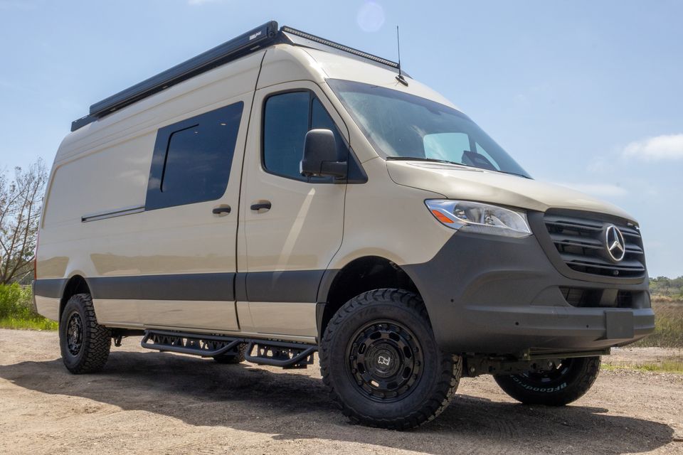 A sand-colored Oasis camper van with black wheels and side steps